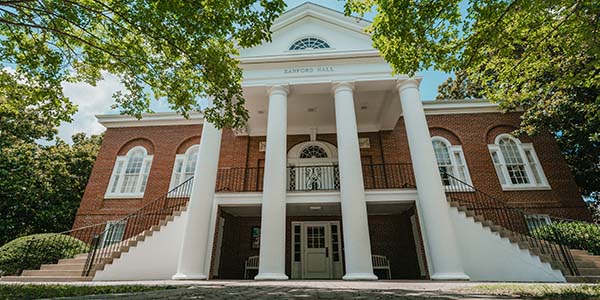 Low-angle exterior view of Sanford Hall, a red brick campus building with large white columns, dual staircase entrances, and surrounding green trees.